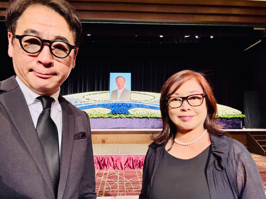 Two people in formal black attire take a selfie on a stage with a blue floral arrangement and a portrait in the background.