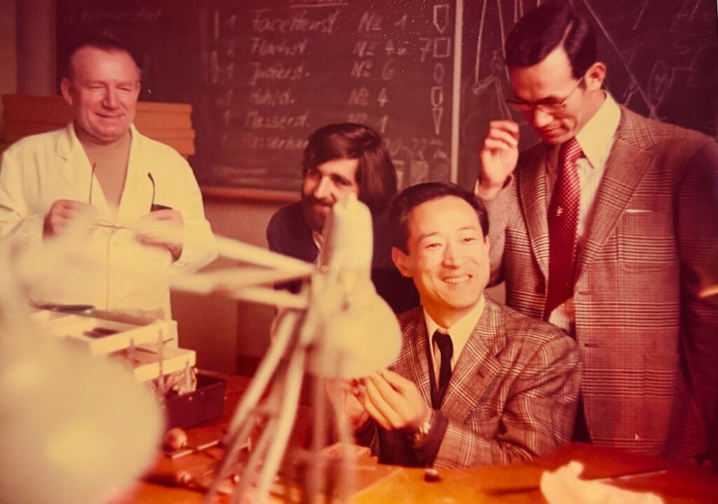 Group of smiling men in a workshop or classroom, with a chalkboard in the background and stacks of papers on the table.