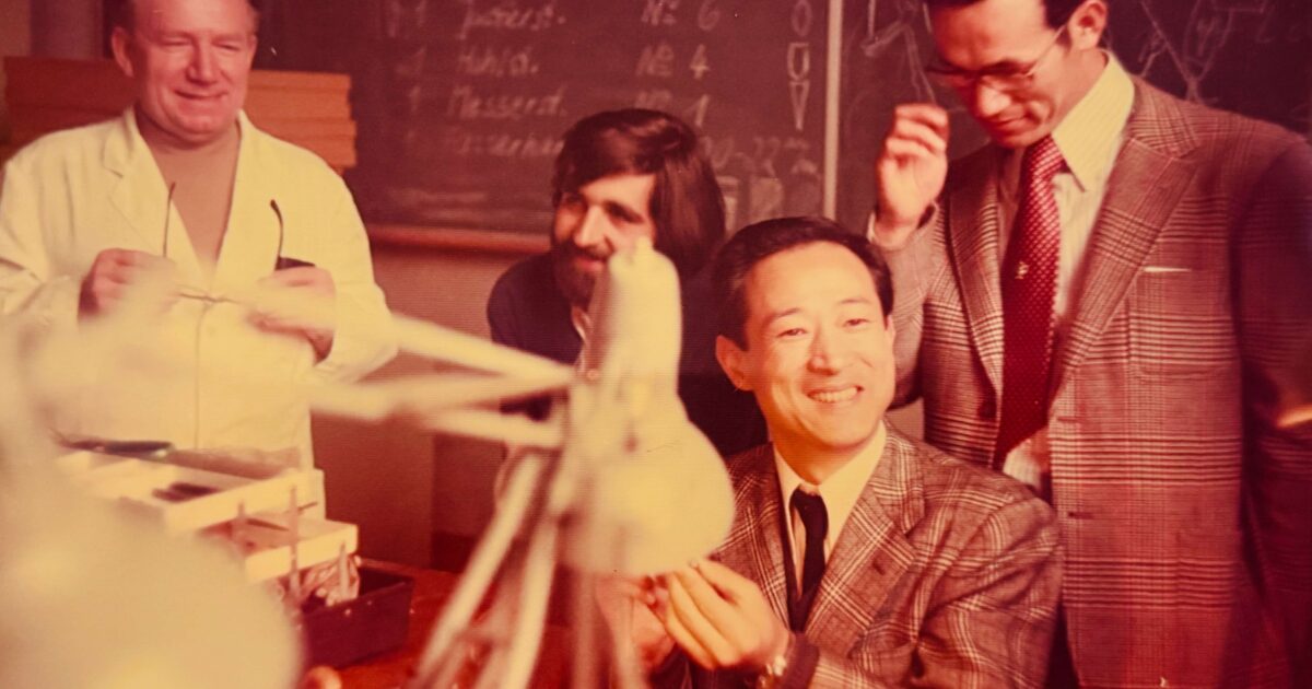 Group of smiling men in a workshop or classroom, with a chalkboard in the background and stacks of papers on the table.
