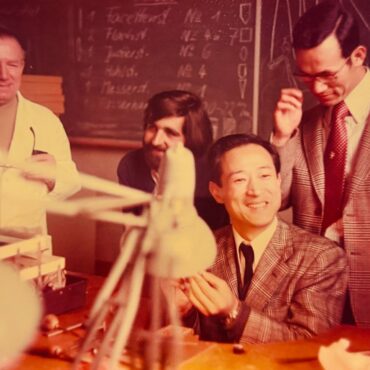 Group of smiling men in a workshop or classroom, with a chalkboard in the background and stacks of papers on the table.
